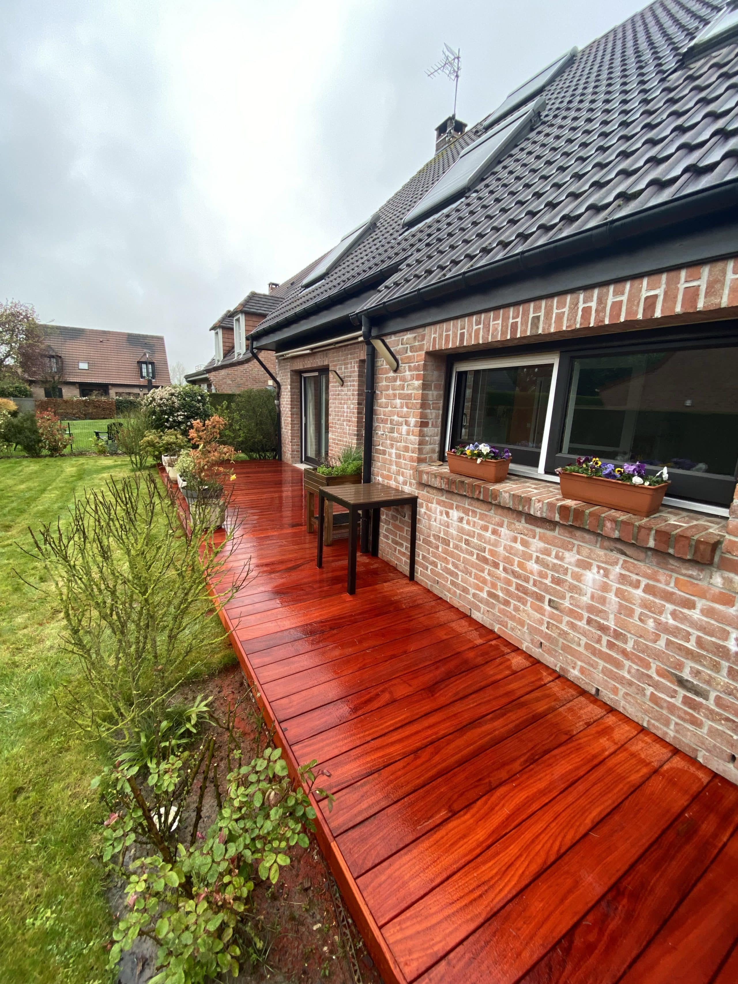 Terrasse en bois rouge vif mouillée par temps humide, avec jardin et maison en arrière-plan
